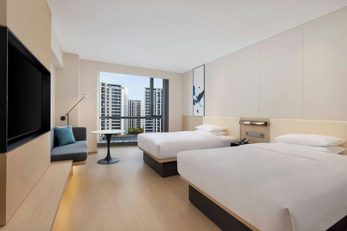 A modern hotel room with two beds, a sofa, a TV, and a large window with a city view.