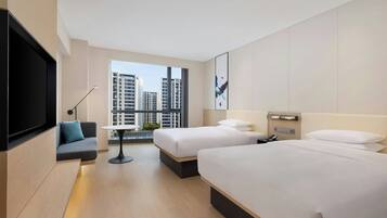 A modern hotel room with two beds, a sofa, a TV, and a large window with a city view.