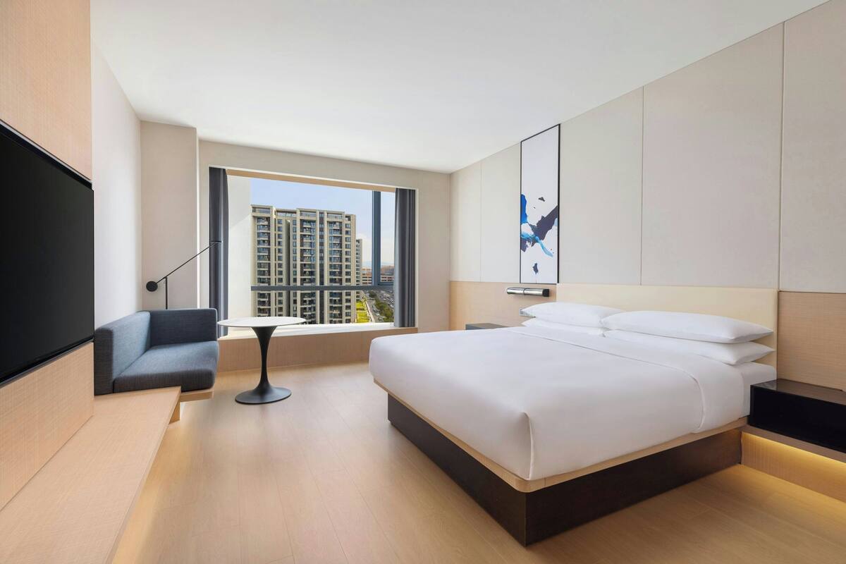 A modern hotel room with a large bed, a seating area, a TV, and a view of the cityscape.