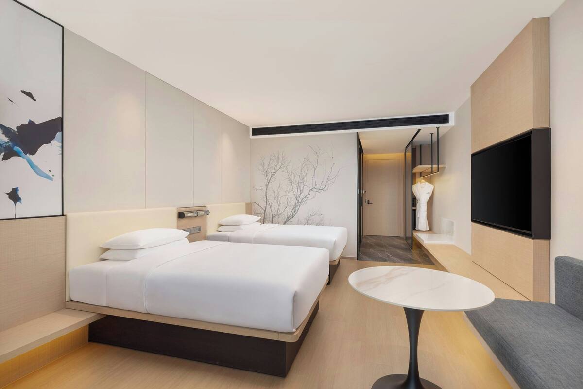 A modern hotel room with two beds, a flat-screen TV, a small round table, and a minimalist decor.