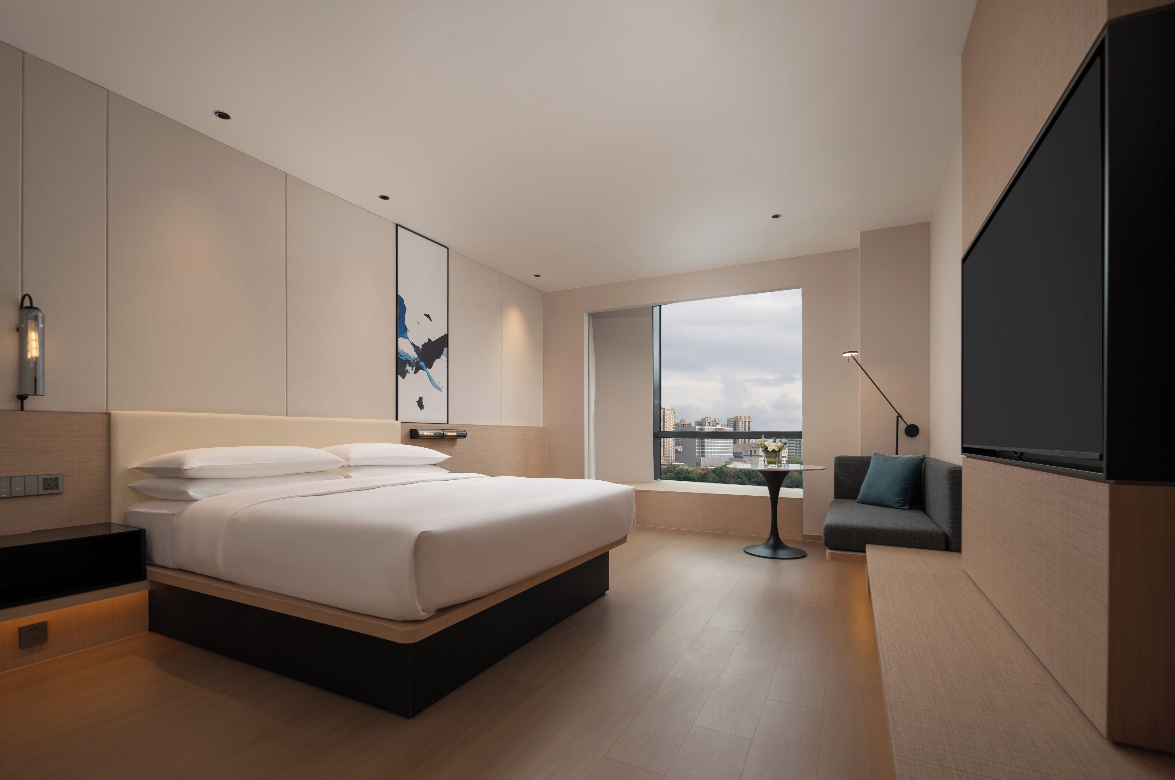 A modern hotel room with a large bed, a seating area with a table, and a large window offering a city view.