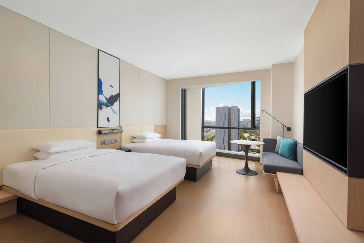 A modern hotel room with two beds, a sofa, a TV, and a large window offering a city view.
