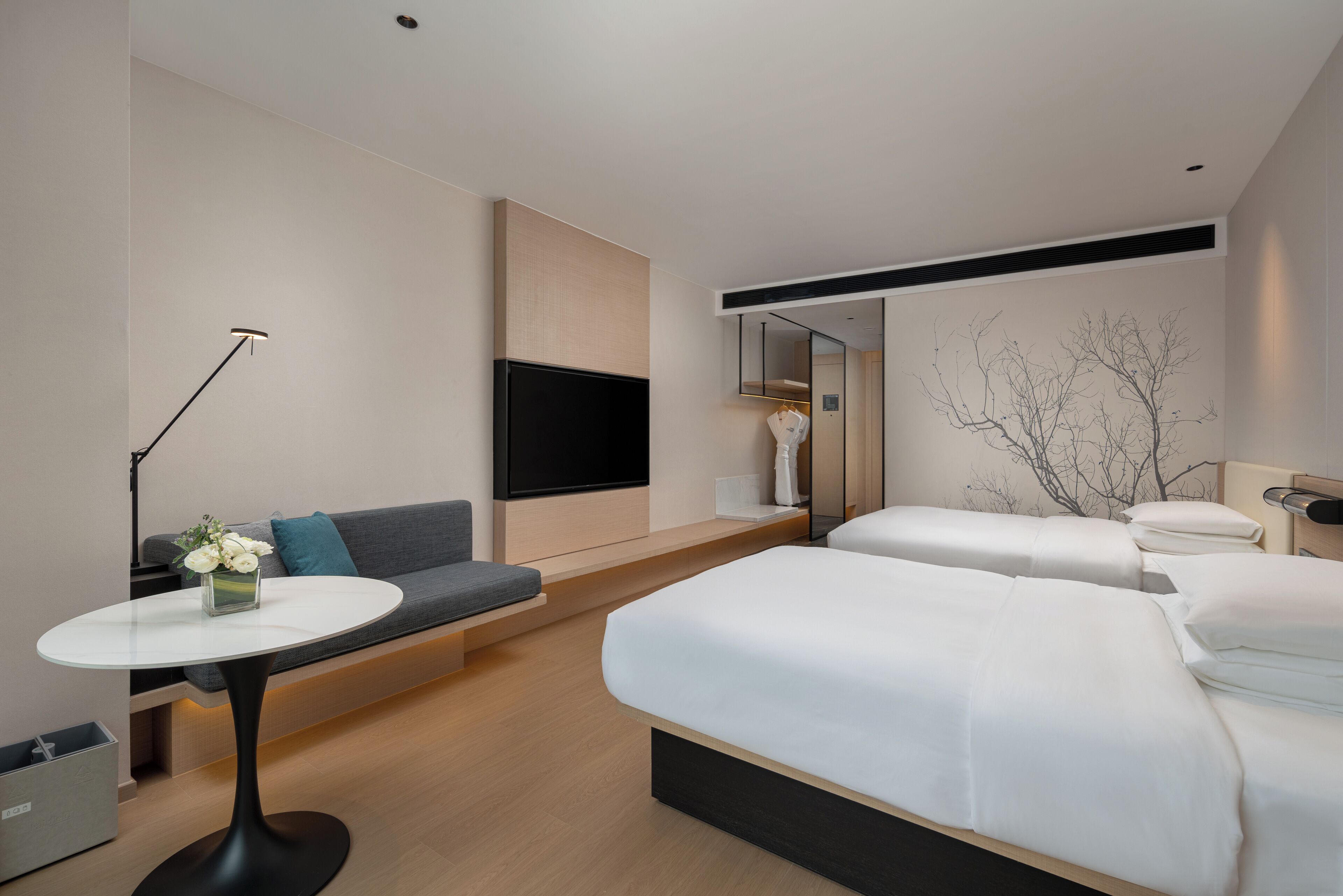 A modern hotel room with a sofa, two beds, a flat-screen TV, a small table with a vase of flowers, and a mirror.