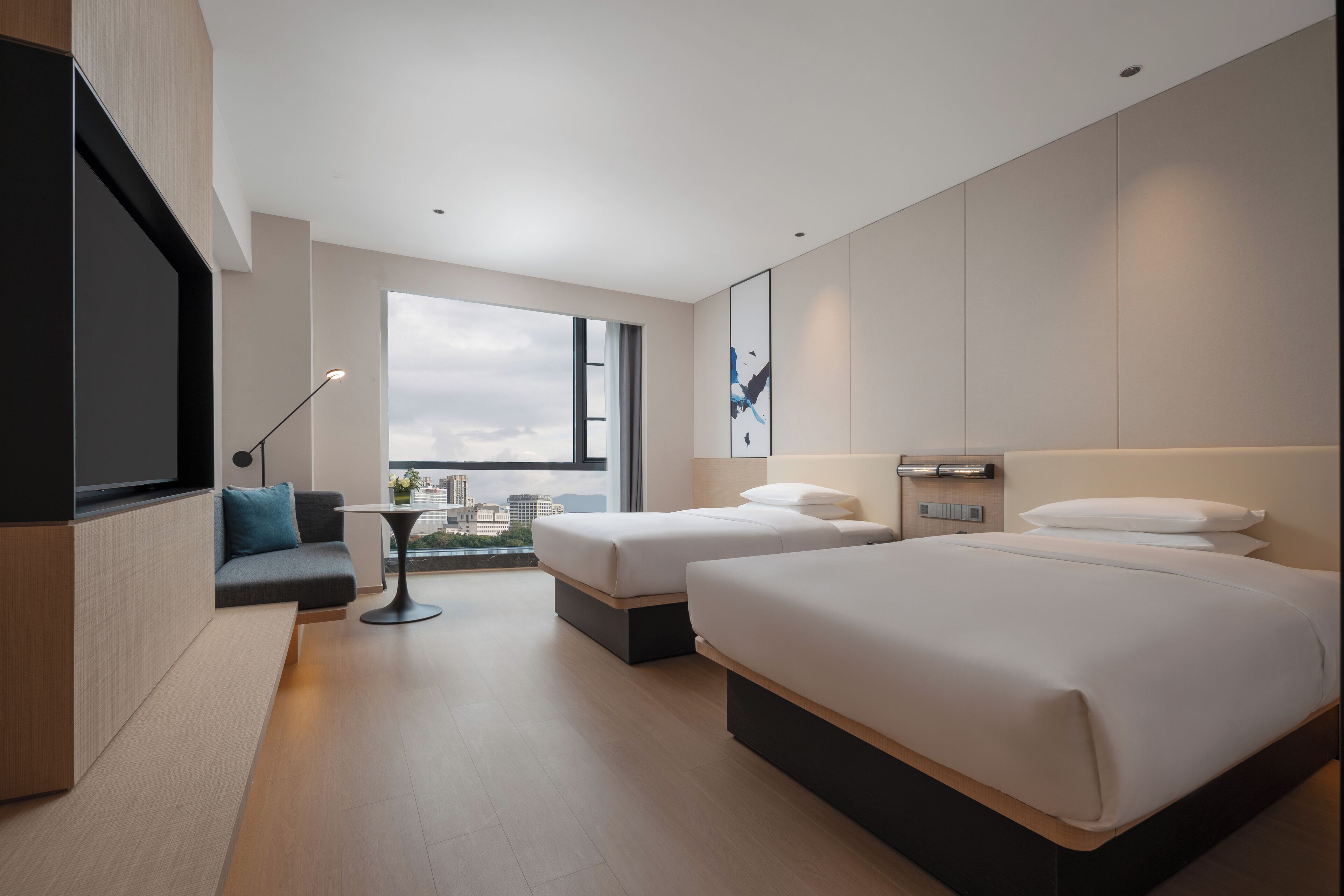 Quarto de hotel moderno com duas camas, um sofá, uma mesa pequena e uma grande janela com vista para a cidade.