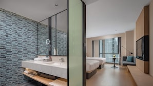Una habitación de hotel moderna con un espejo grande, un baño con salpicadero de azulejos, un sofá, un televisor y un ventanal con vistas a la ciudad.