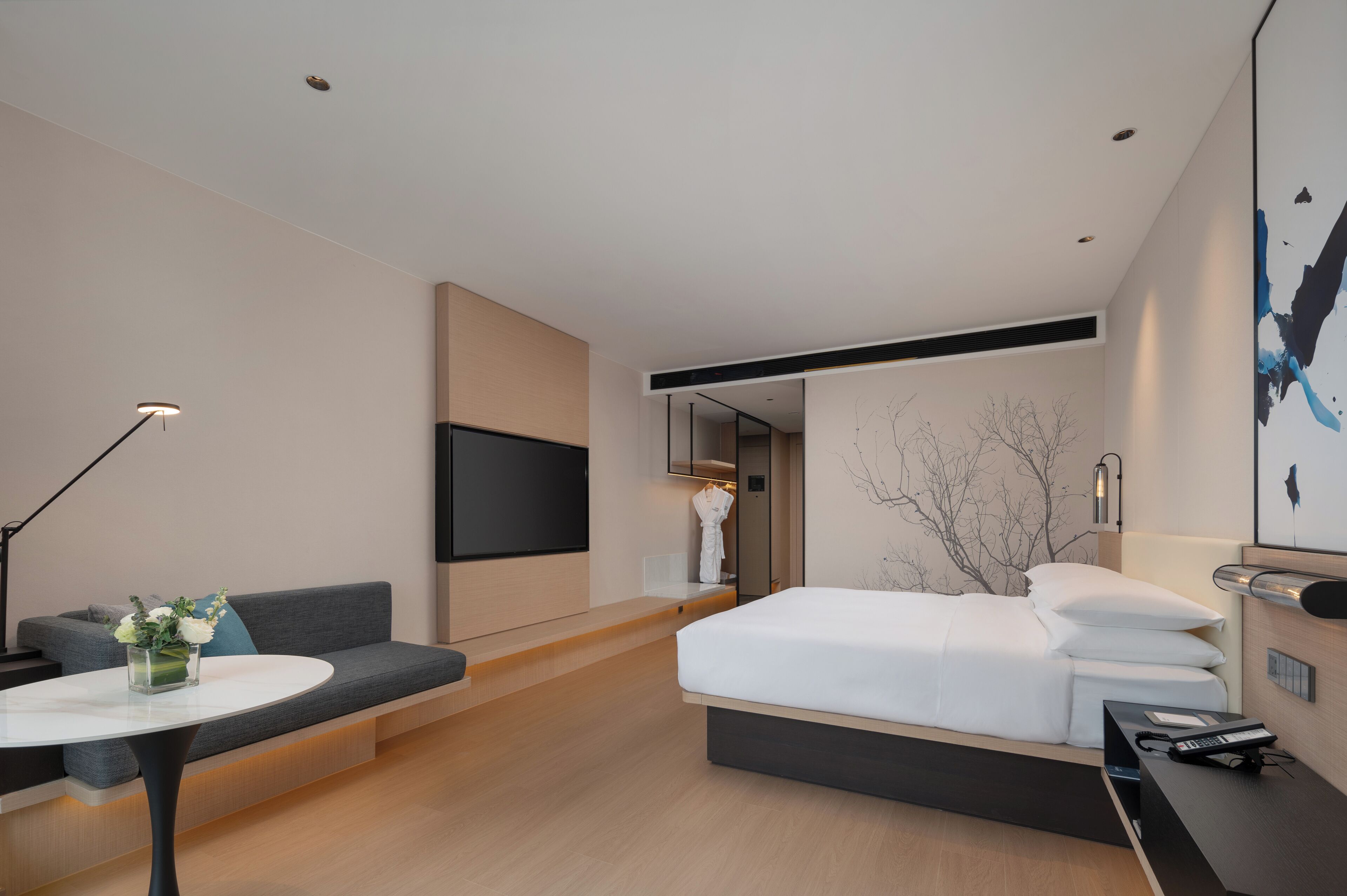 Ein modernes Hotelzimmer mit einer Couch, einem Bett, einem kleinen Tisch mit Blumenvase, einem Flachbildfernseher und einem Gemälde an der Wand.