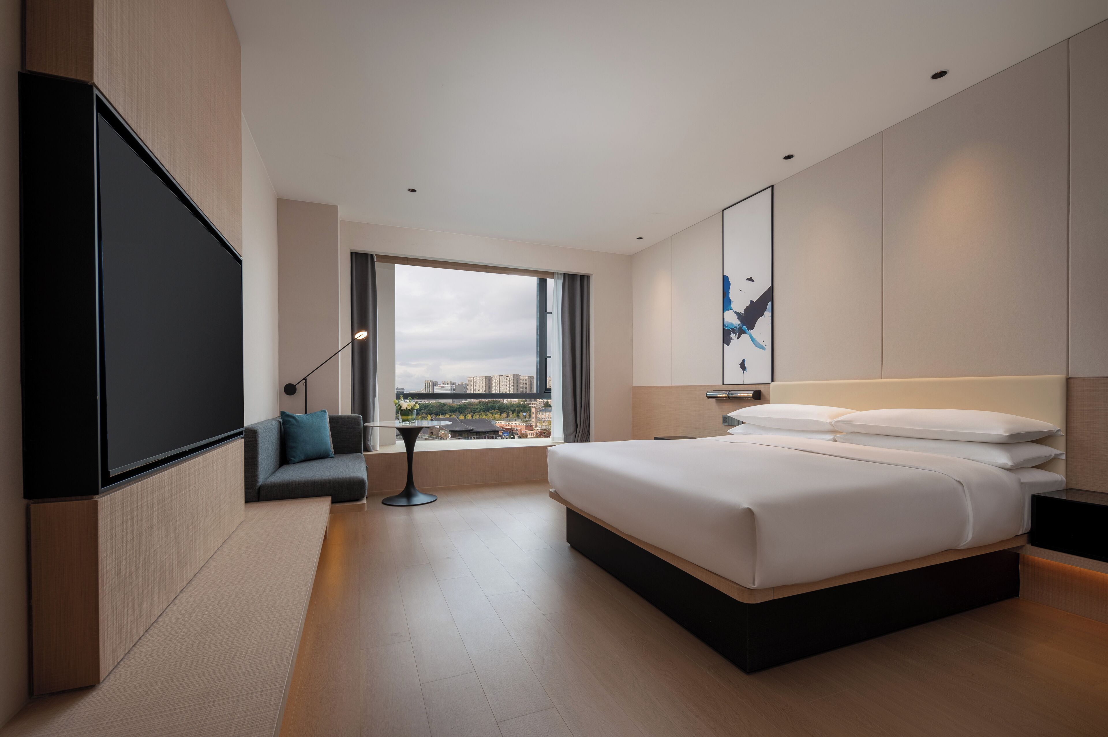 A modern hotel room with a large bed, a seating area with a table, a TV, and a large window offering a city view.