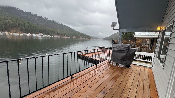 Une terrasse en bois avec une rambarde en métal donnant sur un lac, un barbecue couvert et un bâtiment à l’extérieur blanc.