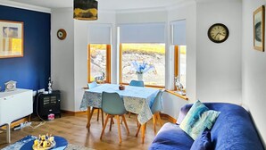 Ein Wohnzimmer mit blauem Sofa, einem Esstisch mit Stühlen, einem kleinen Beistelltisch und Blick auf eine felsige Landschaft durch große Fenster.