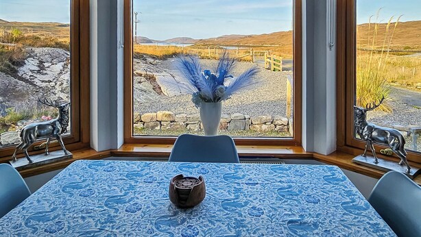 Ein Essbereich mit einem Tisch für zwei Personen, einer Vase mit blauen Blumen und Blick auf eine ländliche Landschaft durch große Fenster.
