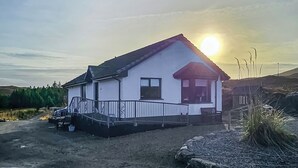 Ein eingeschossiges Haus mit weißer Fassade und schwarzem Dach, auf unbefestigtem Boden gelegen, mit einem Kiesweg, der zum Eingang führt.