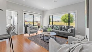 Un salon moderne avec un canapé gris, deux fauteuils, une table basse et une table à manger. De grandes fenêtres offrent une vue sur l’extérieur.