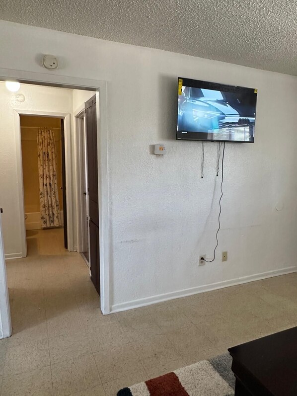 A living room with a flat-screen TV mounted on the wall, a door leading to another room, and a patterned rug on the floor.