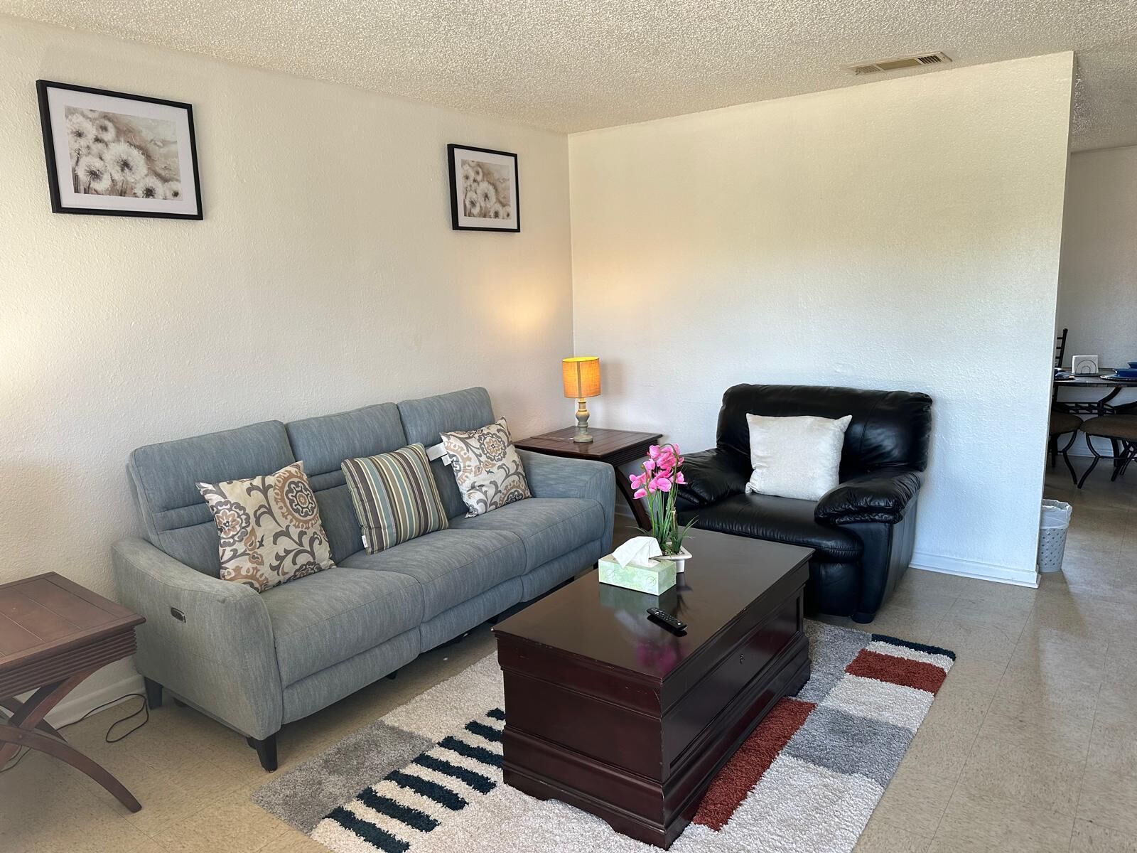 A living room with a grey sofa, a black leather chair, a wooden coffee table with a vase of flowers, and a striped rug.
