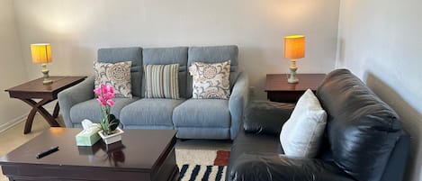 Un salon avec un canapé gris, un divan en cuir noir, une table basse en bois et deux petites tables d’appoint avec des lampes.