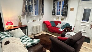 A cozy living room with a brown sofa, a red sofa, and a brown armchair. There is a window with curtains, a radiator, and a small table with a lamp.