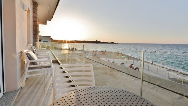 A balcony with white chairs and a table overlooking a beach with people and boats. - DOMO 101 - Deluxe Junior Suite with Sea View (Aglientu)