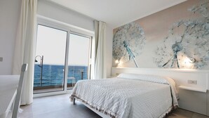 A bedroom with a large bed, a view of the sea through a sliding glass door, and a dandelion artwork on the wall. - DOMO 101 - Deluxe Junior Suite with Sea View (Aglientu)