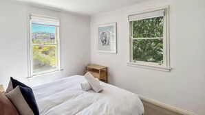Ein Schlafzimmer mit Bett, Kissen, Nachttisch, Lampe und zwei Fenstern mit Blick auf Grünflächen und eine ferne Landschaft.