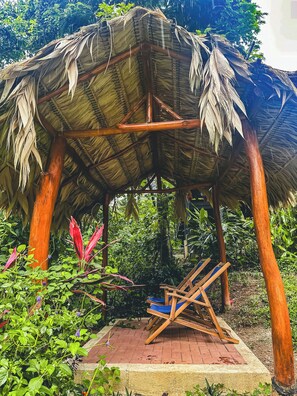 Um gazebo de madeira com telhado de palha, rodeado por vegetação exuberante e um caminho de tijolos.