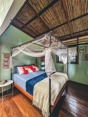 Quarto com cama sob um dossel, uma pequena mesa redonda e um cesto trançado na parede.