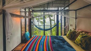 Een kamer met een hangmat, een bed met kleurrijk beddengoed en uitzicht op groen.