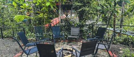 An outdoor seating area with four chairs and a small table, surrounded by lush greenery and a brick pathway.