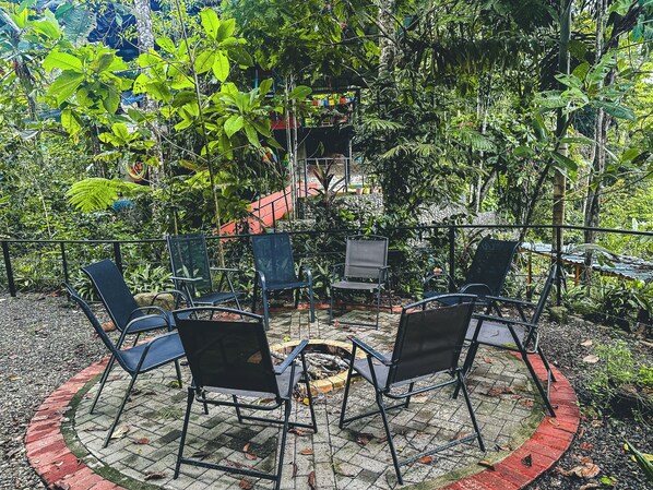 Uma área de estar externa com quatro cadeiras e uma mesa pequena, cercada por vegetação exuberante e um caminho de tijolos.