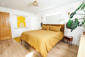 Une chambre avec un plancher en bois, un lit recouvert d’une couvre-lit de couleur moutarde, une porte en bois, une œuvre d’art accrochée au mur, une table de chevet et une plante en pot.