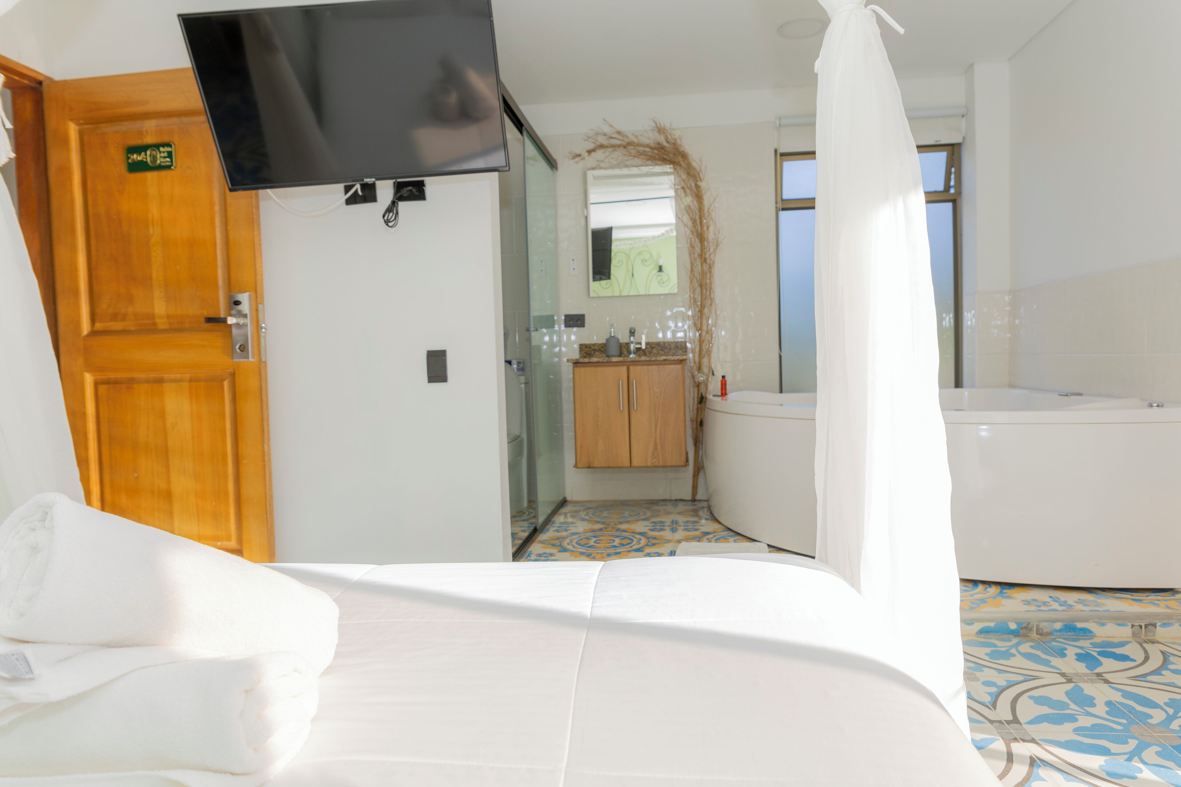 A hotel room with a bed, a TV mounted on the wall, a bathroom with a bathtub, and a small kitchen area.