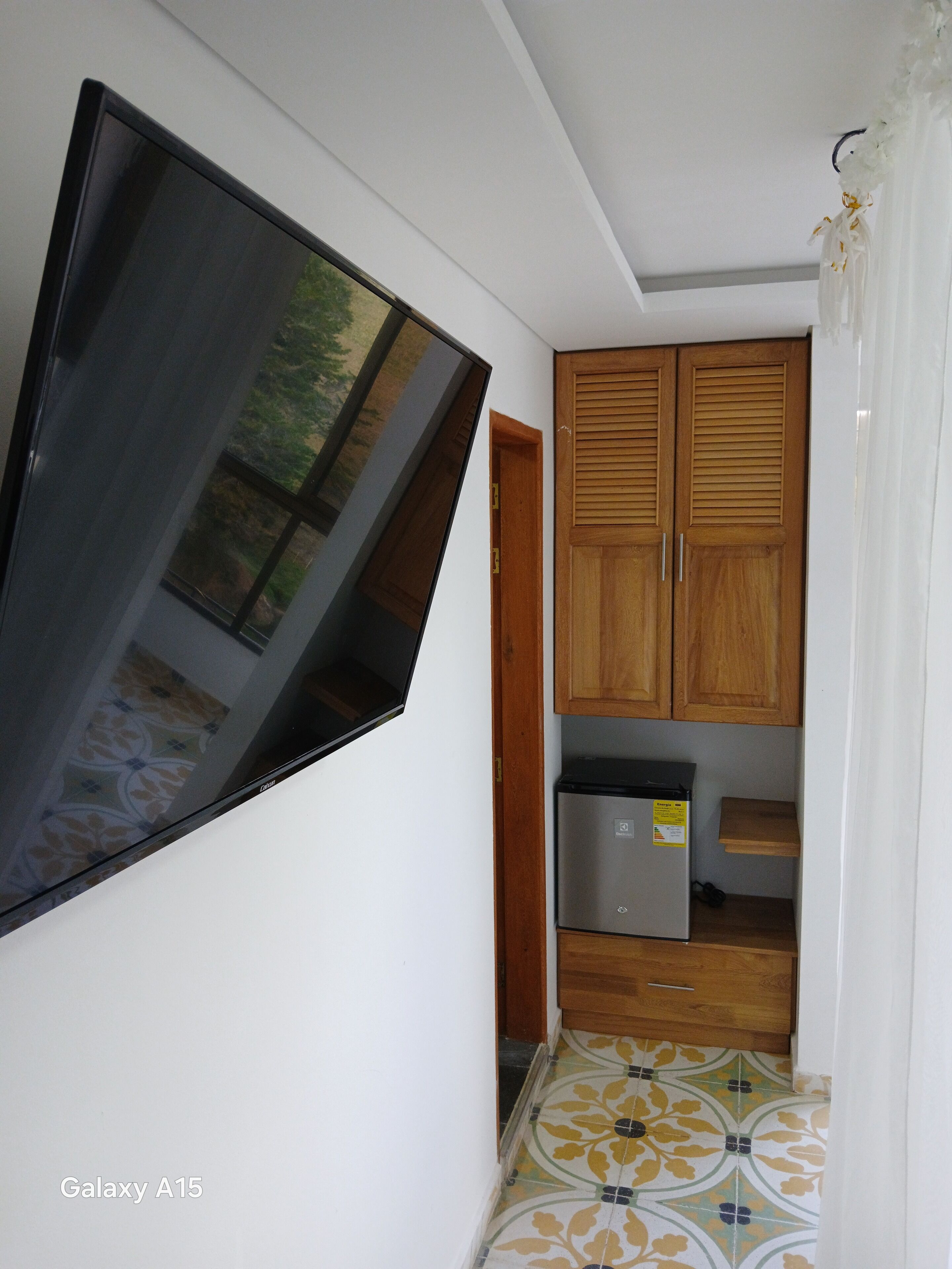 Un corridoio stretto con una grande TV a schermo piatto montata a parete, mobili contenitori in legno, un frigorifero e piastrelle decorative.