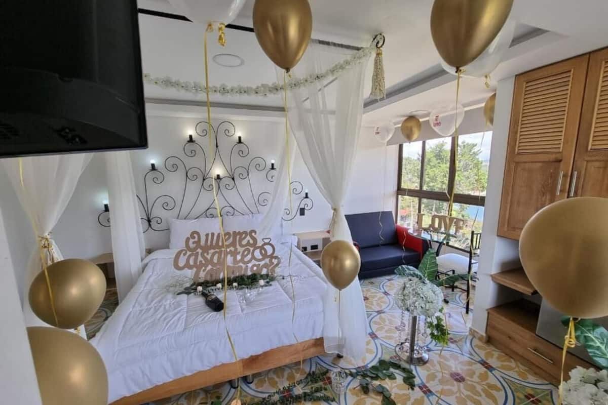 Un dormitorio con cama, dosel blanco, globos dorados y televisor montado en la pared.