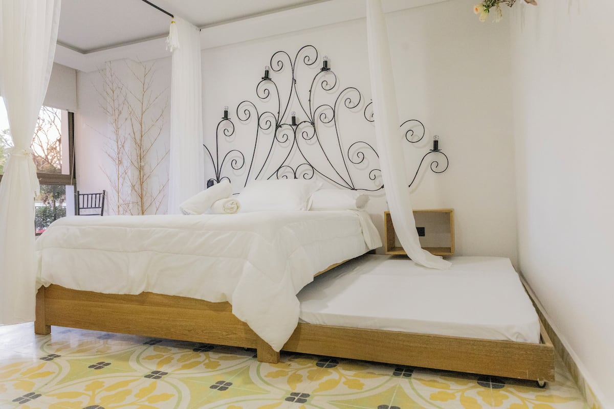 Una cama con estructura de madera y ropa de cama blanca, un cabecero decorativo de metal y una mesita de noche con una lámpara.