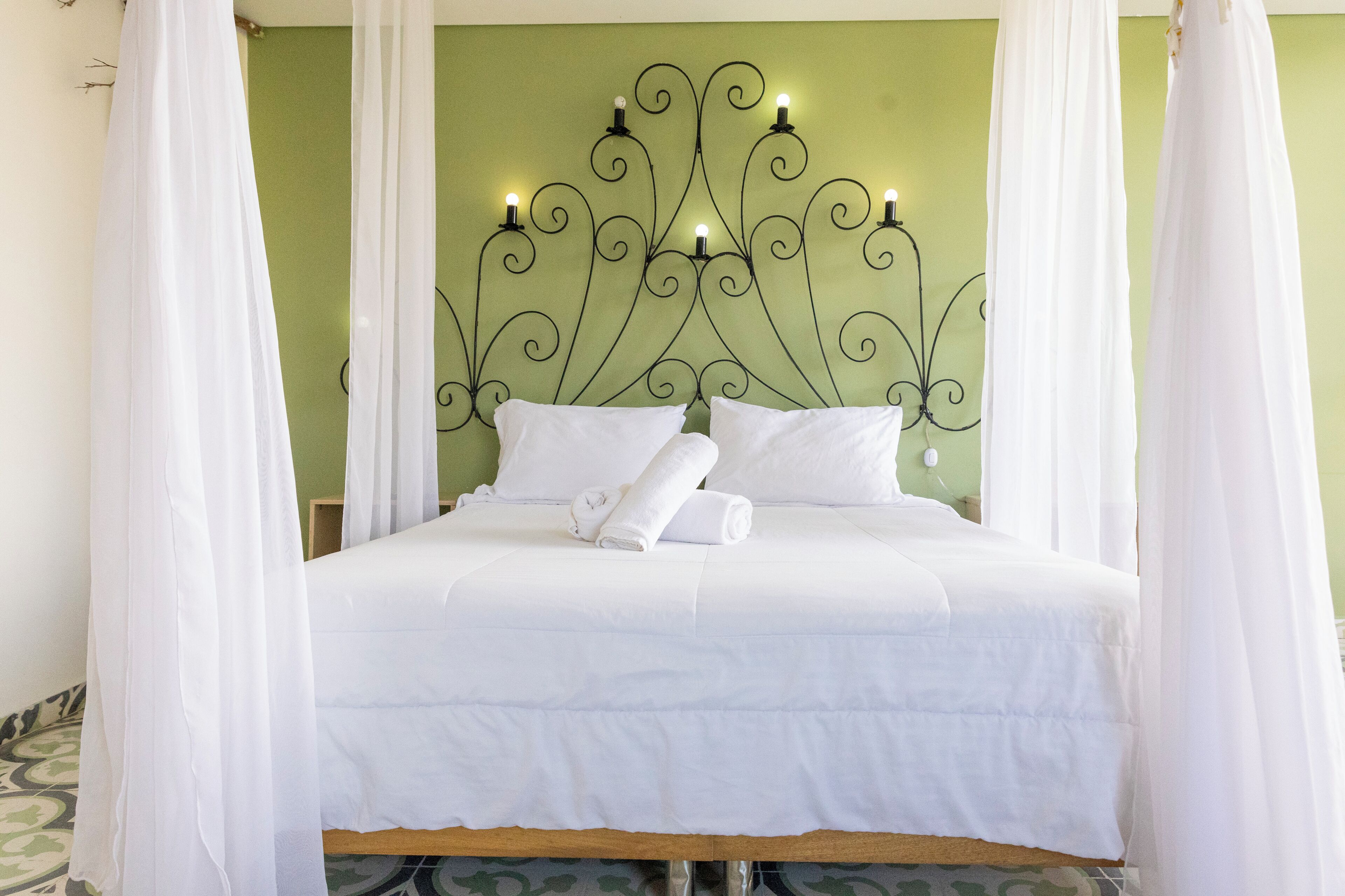 Een keurig opgemaakt bed met witte lakens en kussens, een decoratief metalen hoofdbord met verlichting, en lichte witte gordijnen.