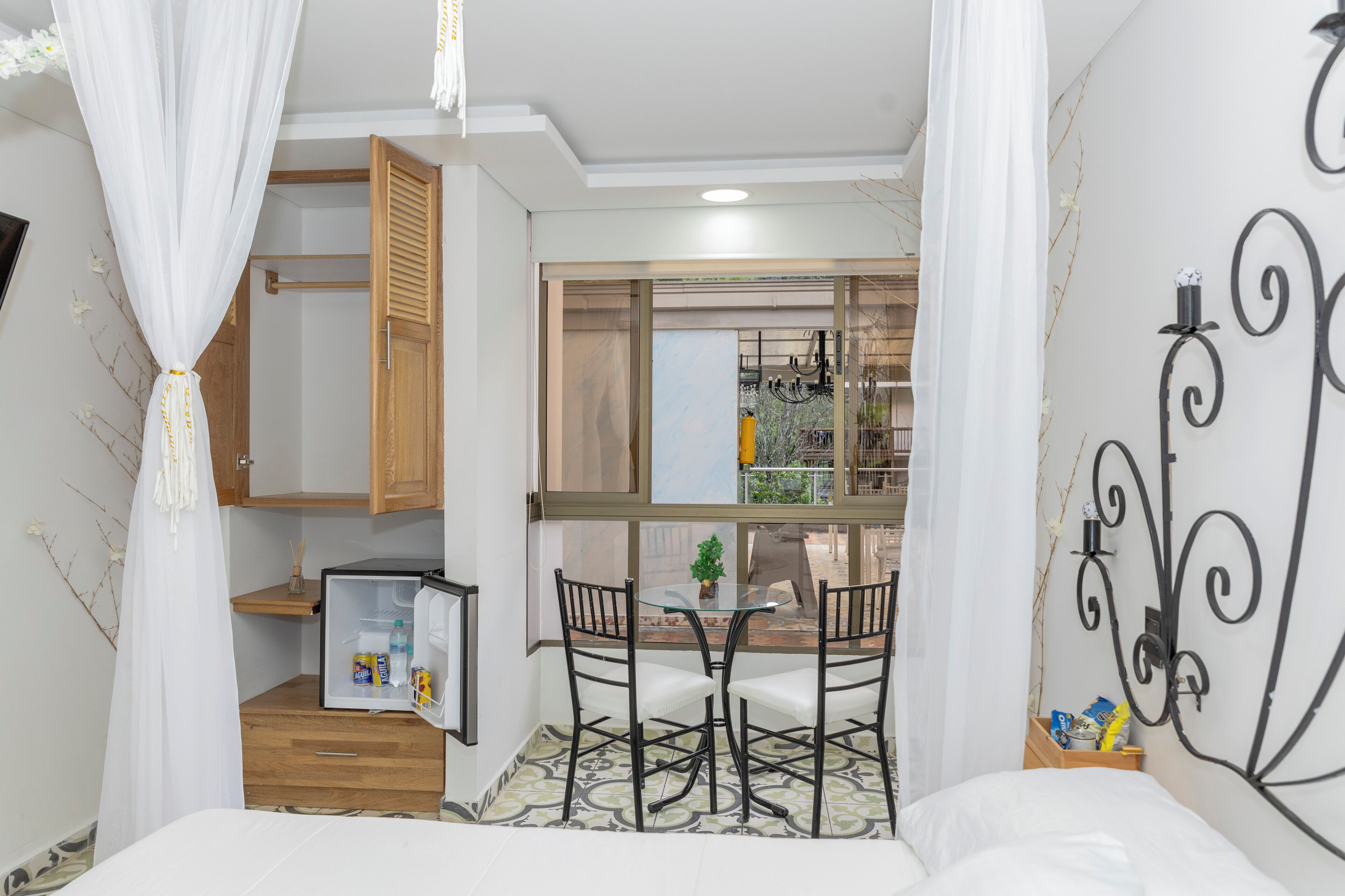 A compact hotel room with a bed, a small dining table with chairs, a refrigerator, and a window with a view of the outside.