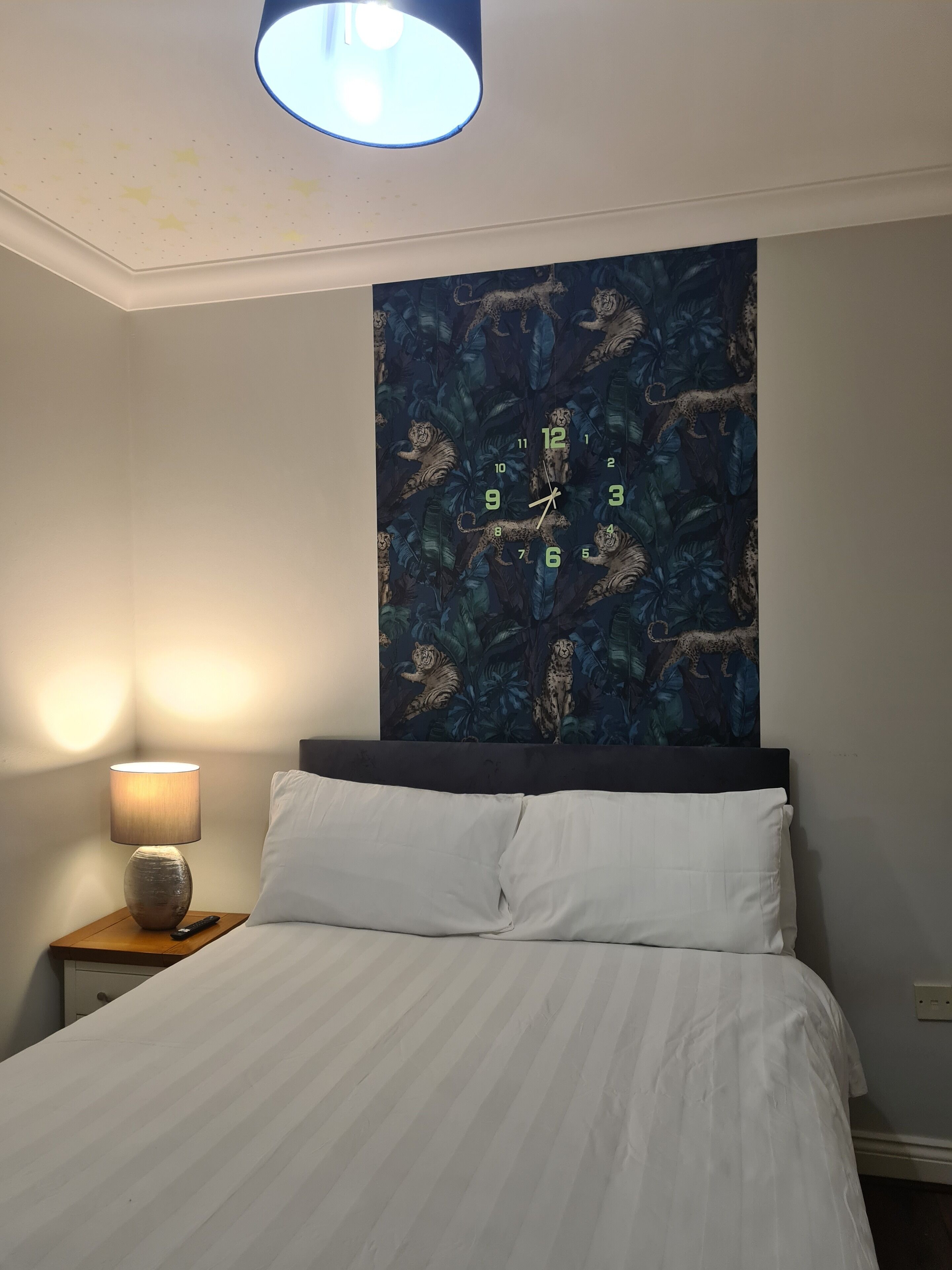 Een slaapkamer met een bed, nachtkastje, lamp en een wandklok met een jungle ontwerp.