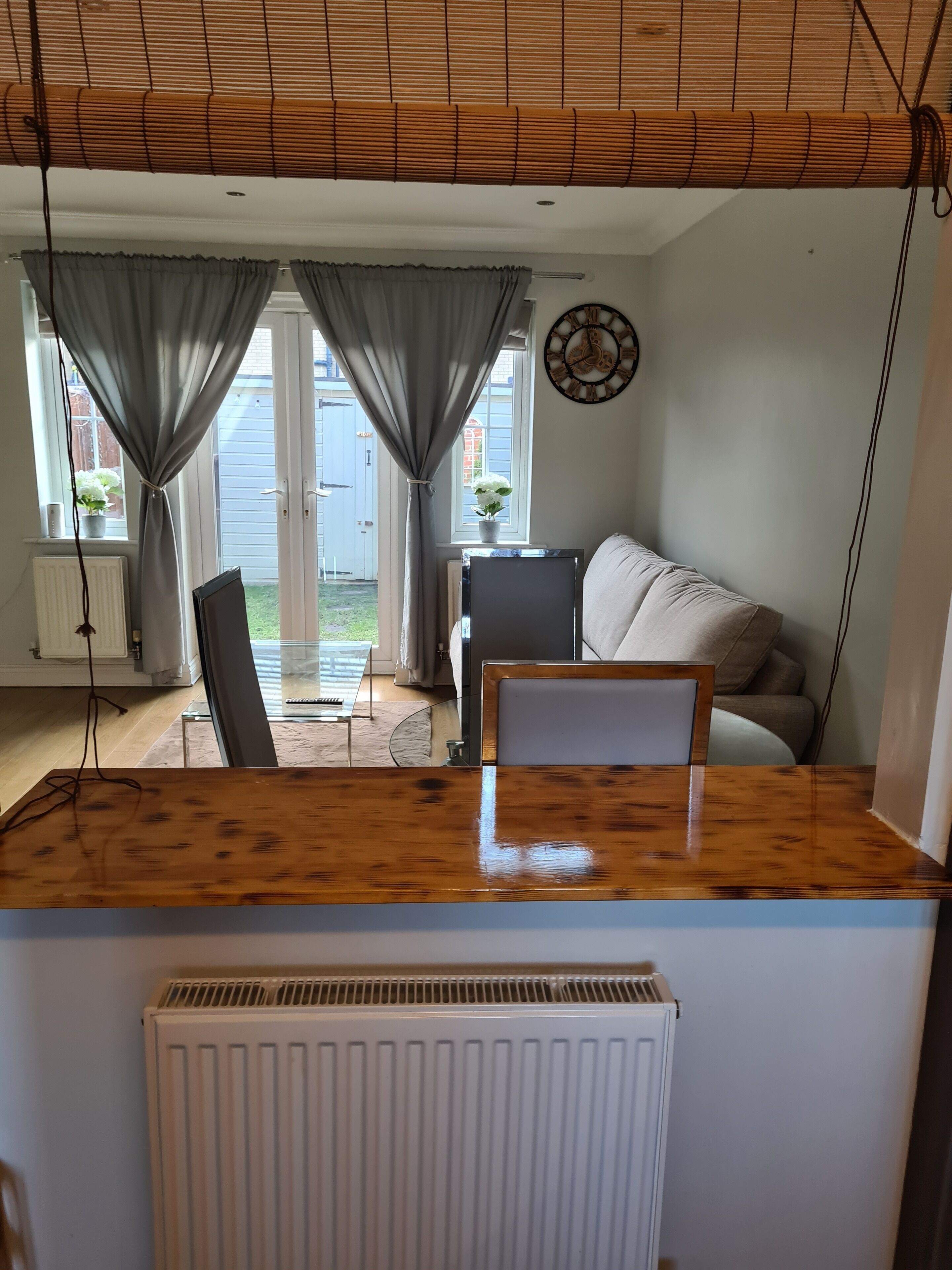 A room with a wooden table, a white radiator, a grey sofa, and a window with curtains.