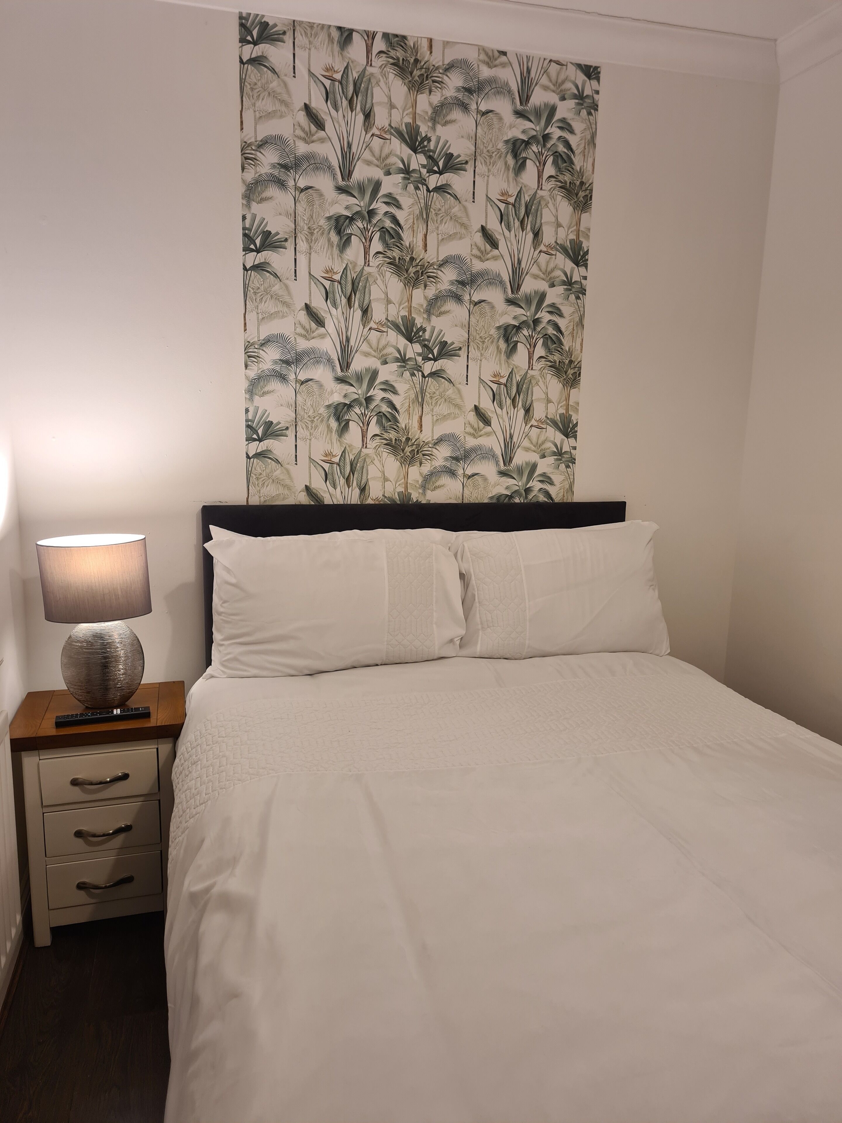 Una cama bien tendida con ropa blanca, una lámpara de noche y una mesita de noche con cajones.