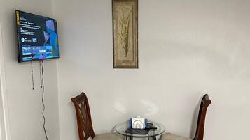 Una habitación con un televisor de pantalla plana en la pared, un cuadro botánico enmarcado, dos sillas de madera, una mesita redonda de vidrio y un enchufe en la pared blanca.