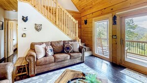 Ein Holzinnenraum mit einer Treppe, einem braunen Sofa mit Tierkissen und Blick ins Freie durch große Fenster.
