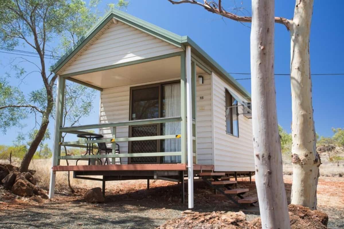 A small, single-story cabin with a porch and a table, surrounded by trees and dry ground.