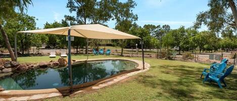 A backyard with a swimming pool, a covered patio area, and blue lawn chairs.