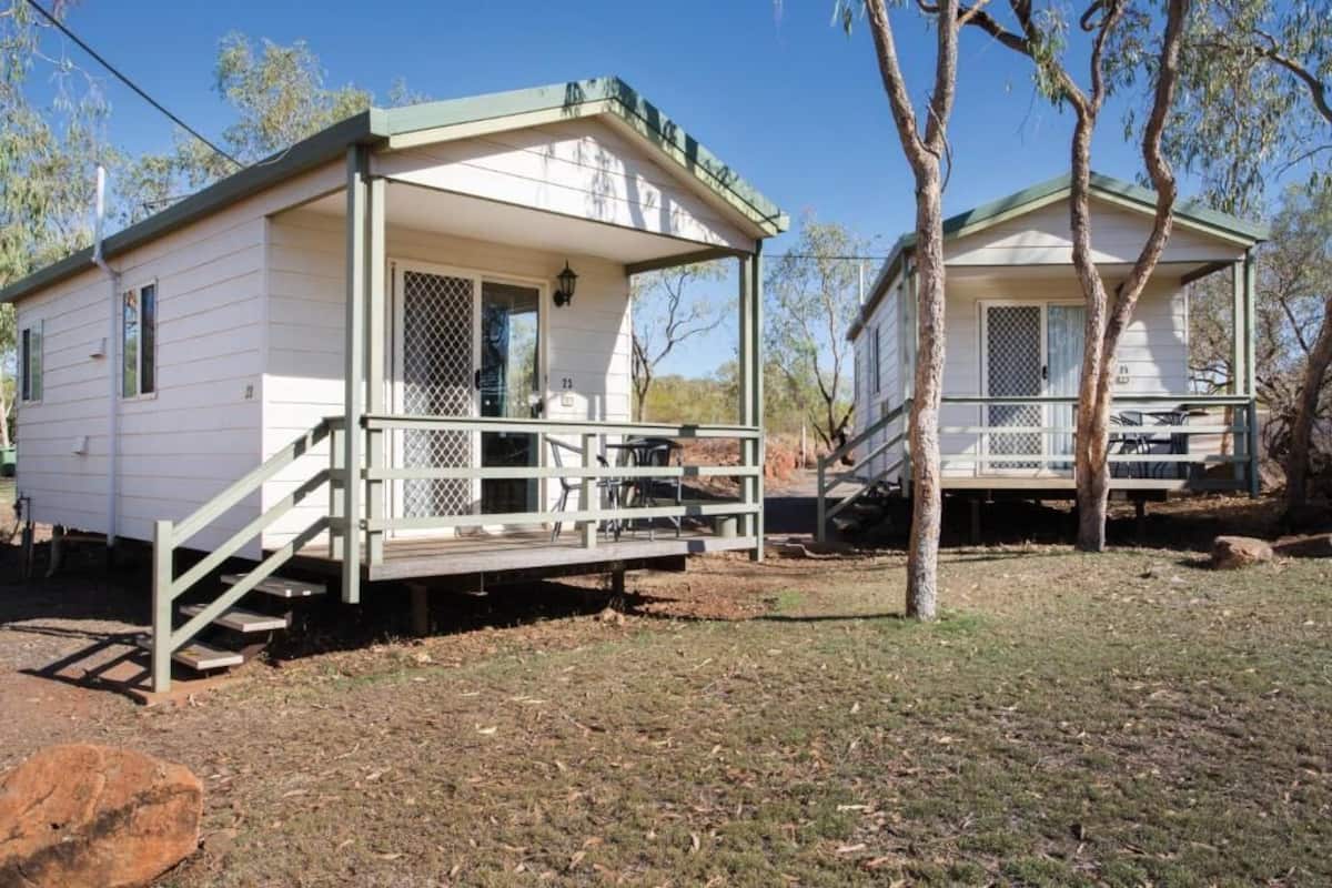 Two small, single-story cabins with a porch and a small table, surrounded by trees and a clear sky.
