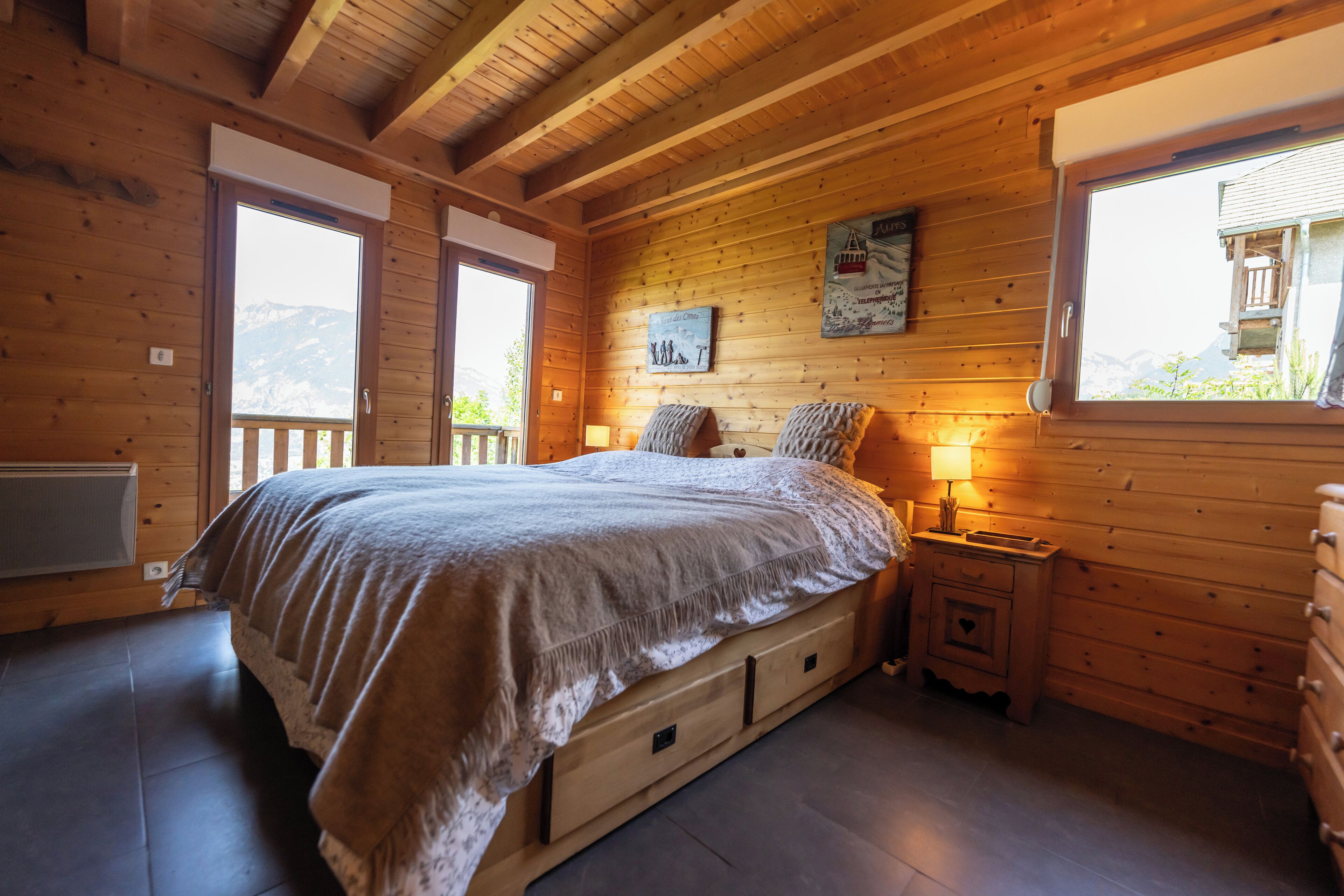 A wooden cabin bedroom with a large bed, a nightstand, a dresser, and a window with a view of mountains.