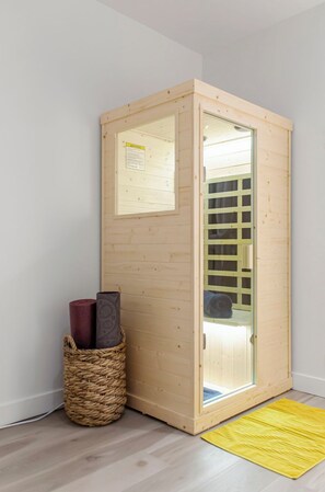 A wooden sauna with a glass door, a woven basket with candles, and a yellow mat on the floor.