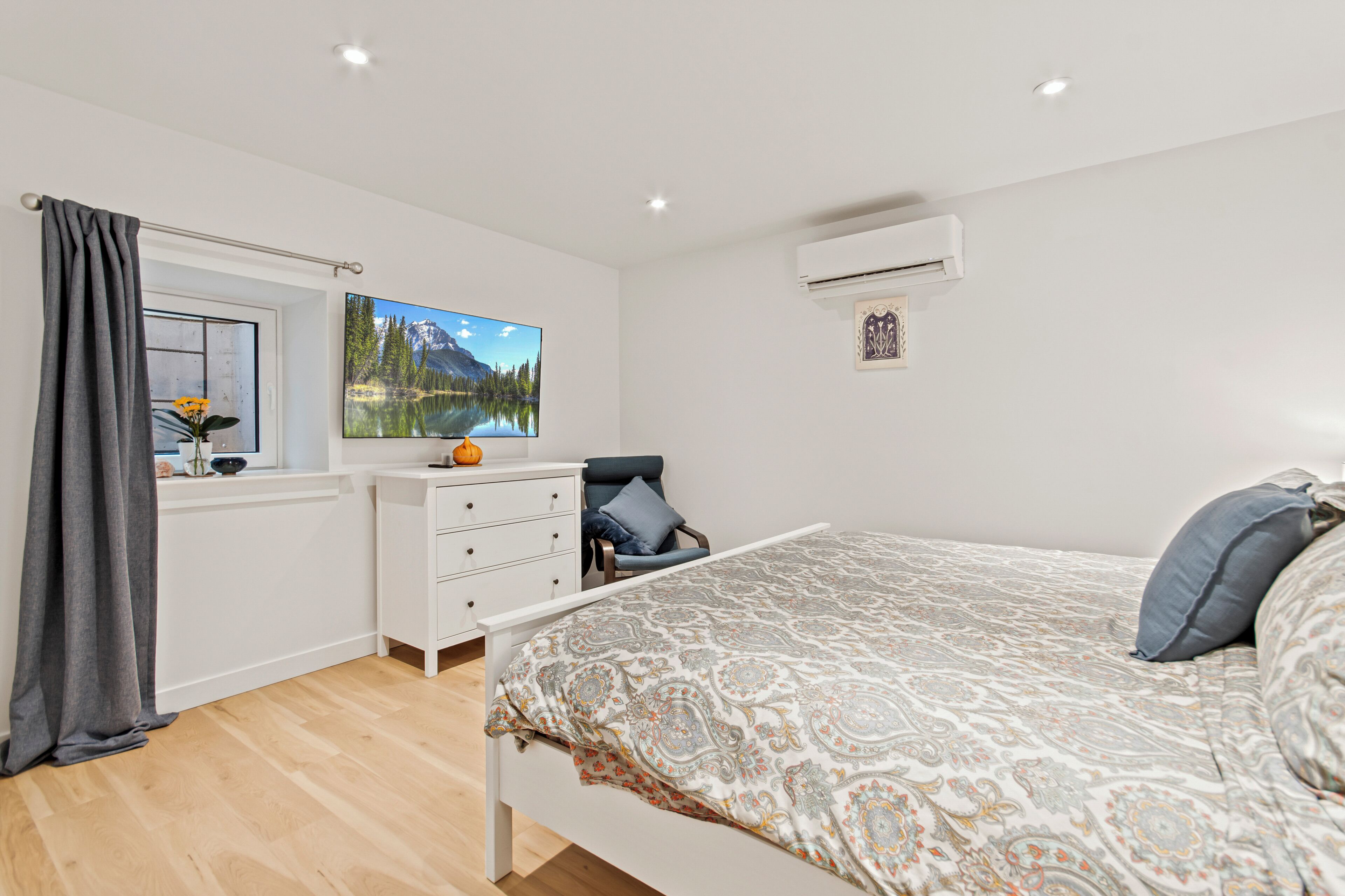A bedroom with a bed, a window with a view of mountains, a dresser, and an air conditioning unit.