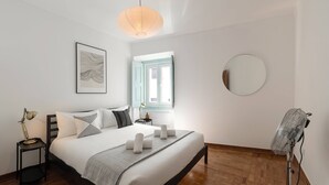 Un dormitorio ordenado con cama, mesitas de noche, lámpara de noche, lámpara de pie, espejo y ventana con vistas.
