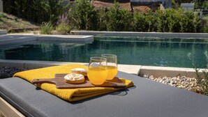 Escena junto a la piscina con una silla de descanso, una toalla amarilla, una tabla de madera con un vaso de jugo de naranja y una galleta.