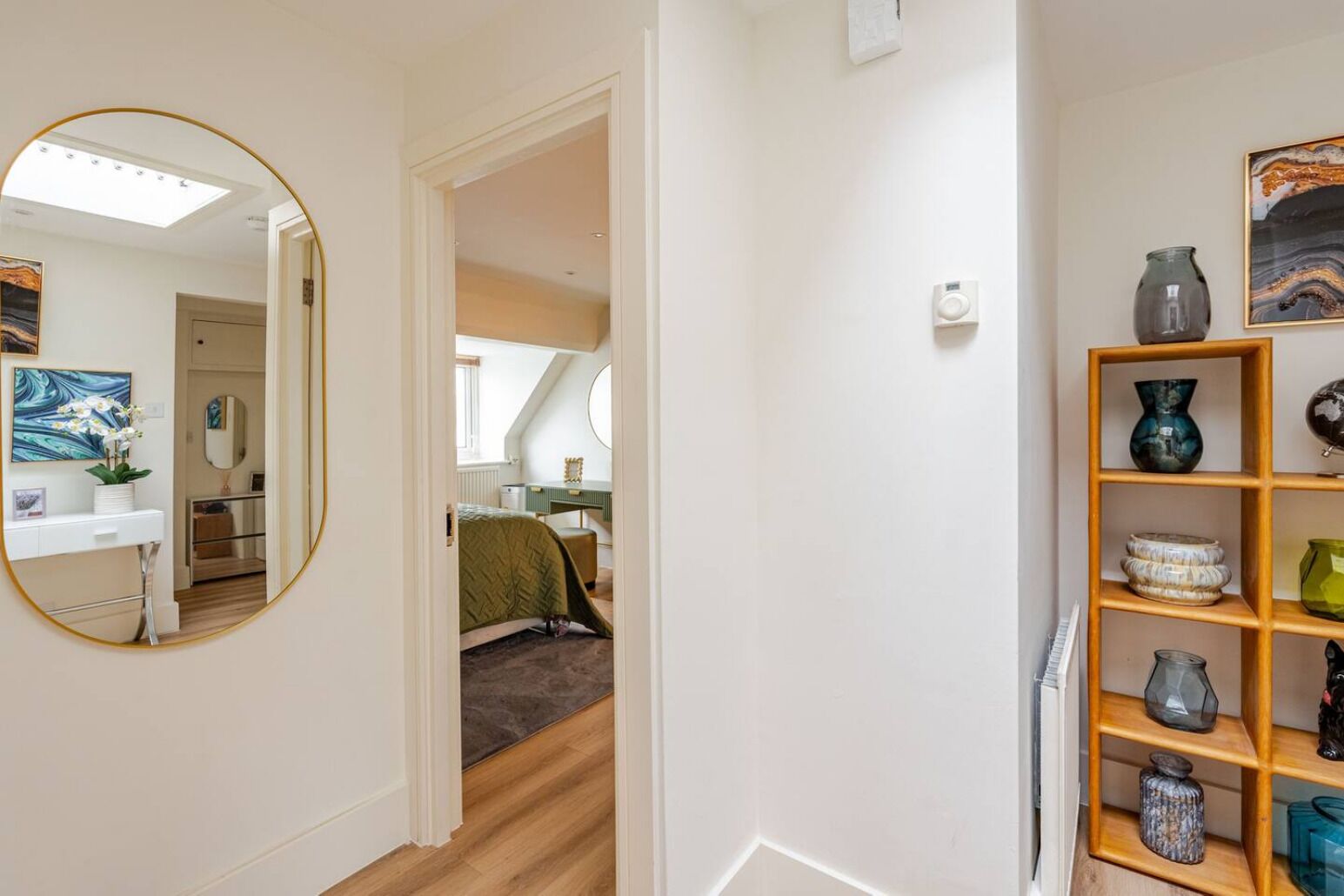 Un couloir avec un miroir, une étagère en bois ornée d’objets décoratifs, et un aperçu d’une chambre avec un lit et un bureau.