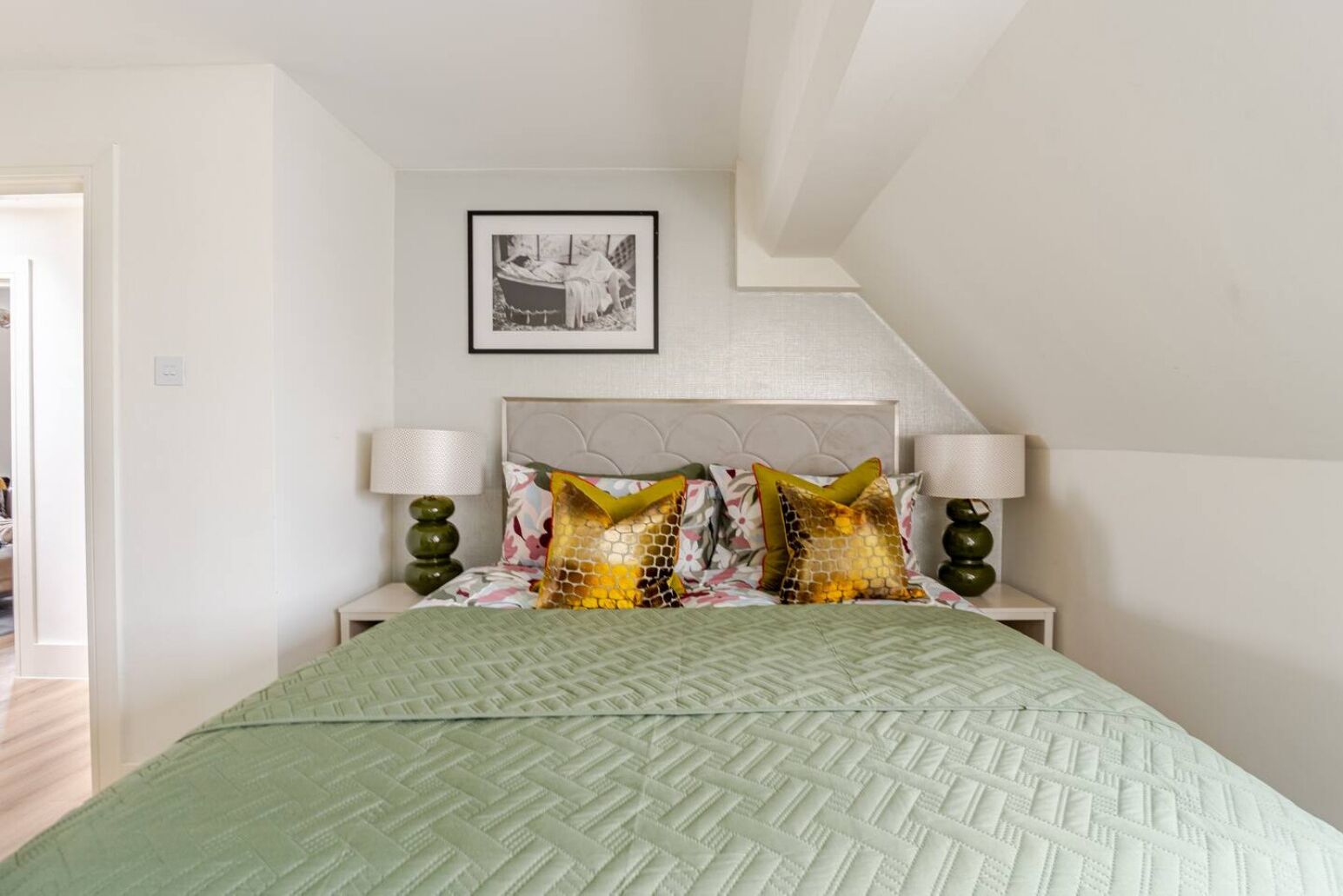 Une chambre à coucher avec un lit, deux lampes de chevet, un tableau encadré au mur et une porte sur le côté gauche.
